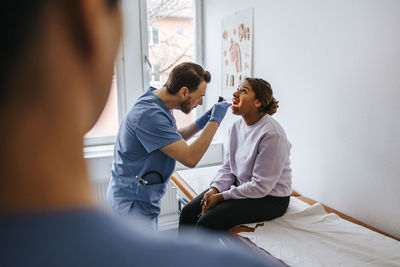 Male doctor examining throat of woman using tongue depressor and flashlight in examination room