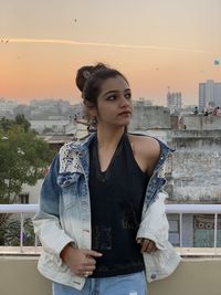 Young woman looking away while standing against cityscape during sunset