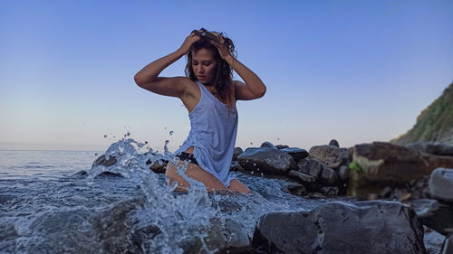 Full length of young woman on rock at beach against clear sky