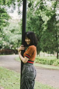 Smiling young woman standing by pole against trees in park
