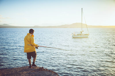 Young man fishing on the coast with a sailing ship in front