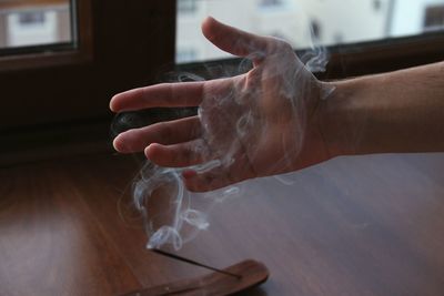 Cropped image of hand touching smoke emitting from incense on table