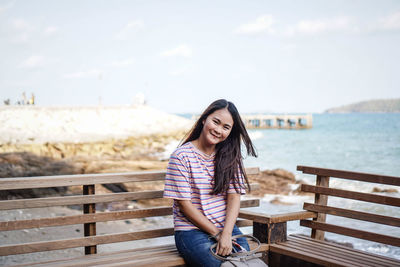 Portrait of smiling young woman sitting on bench by sea