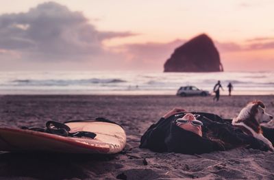 Woman lying with dog by surfboard at beach during sunset