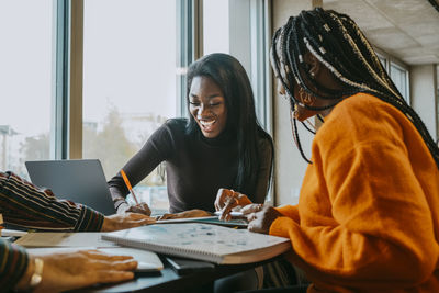 Cheerful female friends studying together while sitting at table in college cafeteria