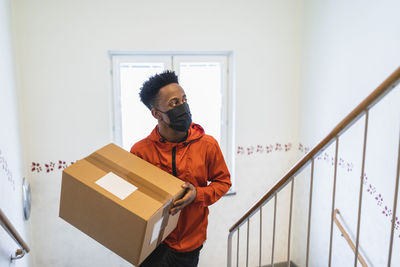 Young delivery man carrying package while moving up on staircase during pandemic