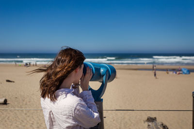 Woman looking through telescope at beach against clear blue sky