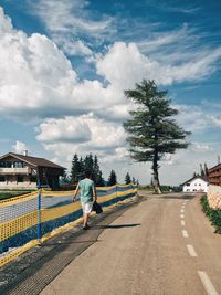 Man walking by fence on roadside in town against cloudy sky