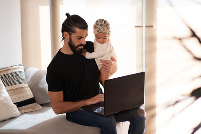 Busy ethnic man sitting on couch with adorable toddler and browsing laptop while working remotely from home