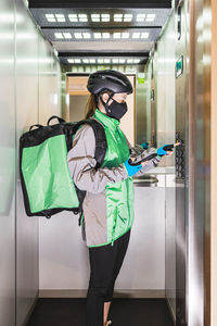 Female courier in uniform and mask using smartphone and pushing button in elevator while making delivery during pandemic