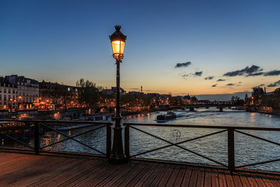 Illuminated street light by river against sky at sunset