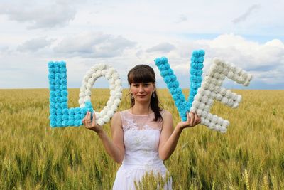 Portrait of beautiful woman holding love text while standing on grassy field against cloudy sky