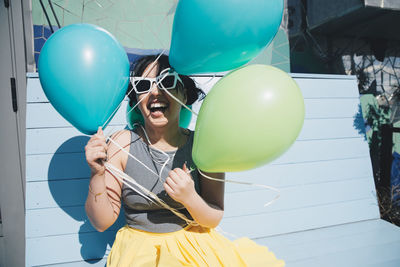 Happy young woman holding balloons while sitting on bench