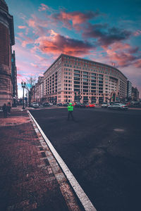 Road by buildings against sky during sunset in city