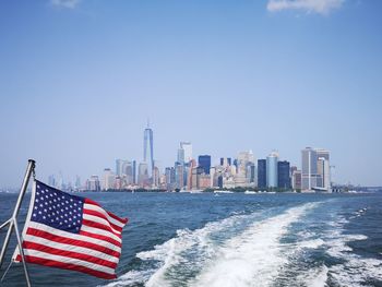 American flag waving against wake in sea with cityscape in background