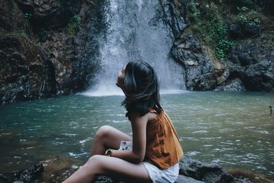 Side view of woman sitting on rock against waterfall