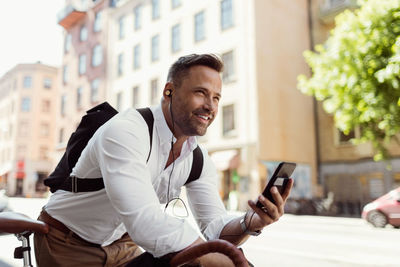 Smiling businessman looking away while holding smart phone on street