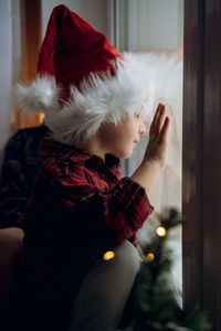 Cute caucasian child wearing santa hat and plaid shirt sitting by the window waiting for christmas