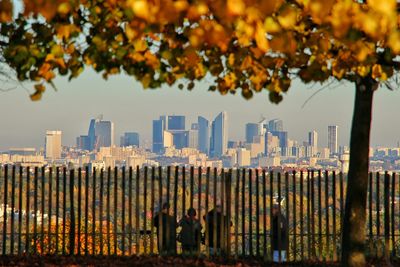 Rear view of people standing by fence against cityscape during autumn