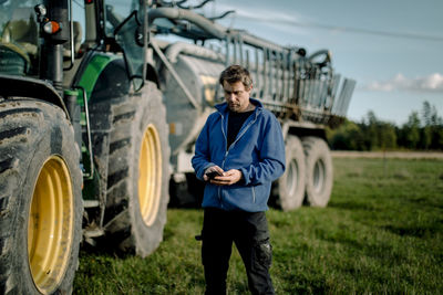 Mature farmer using smart phone standing against tractor on field