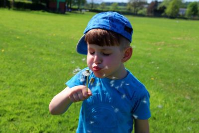 Close-up of cute boy blowing dandelion seed while standing on grassy field at park