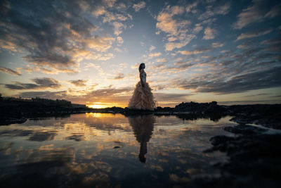 Side view of woman standing by sea against sky during sunset