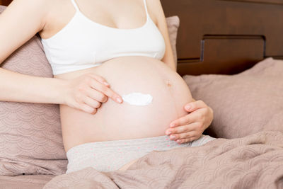 A pregnant girl sits at home on the bed and smears an anti-stretch mark cream on her stomach.