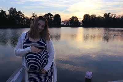 Portrait of pregnant woman touching abdomen while standing by lake against sky during sunset