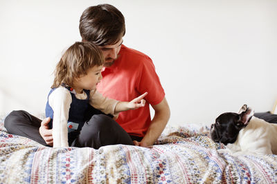 Girl pointing at dog while sitting with father on bed