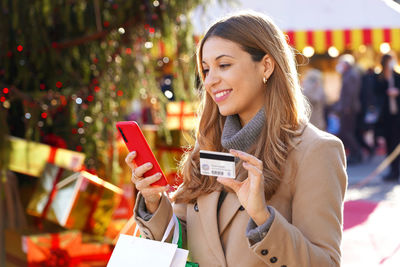 Beautiful smiling girl paying her gifts with mobile phone with christmas markets in the background.