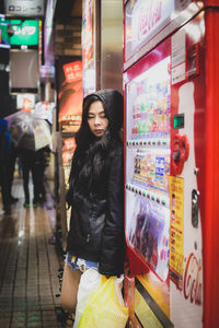 Mid adult woman wearing raincoat standing by display cabinet in city at night