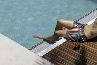 Unrecognizable young man on swimwear in the wooden area with palm tree shadow and a drink glass