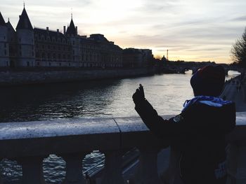 Silhouette woman photographing by river in city against sky during sunset