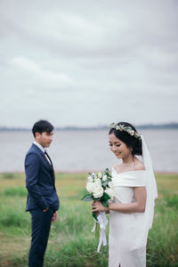 Portrait of bride holding bouquet standing with groom against lake