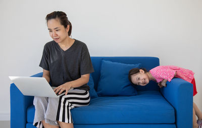 Girl playing with mother using laptop on sofa against wall