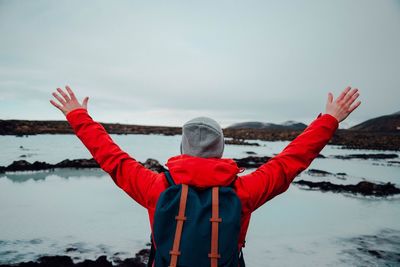Rear view of hiker with arms raised standing by lake against sky