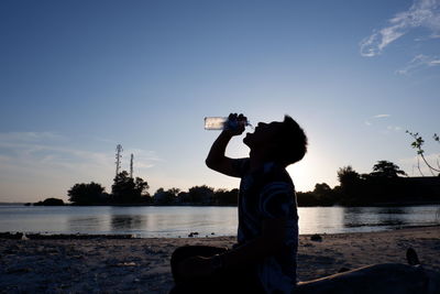 Rear view of man with arms raised standing by lake against sky during sunset