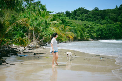 Side view of mid adult woman with dog walking at beach against trees