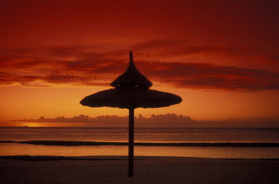 Silhouette thatched roof on beach against orange sky