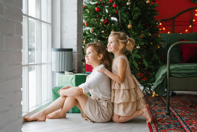 Girls children sit at the window against the background of a christmas tree in the bedroom, look