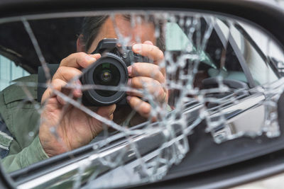 Reflection of man photographing on shattered side-view mirror