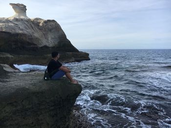 Full length of young man sitting on rock formation at beach against sky