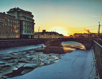 Bridge over river amidst buildings against sky during sunset