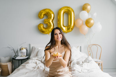 Positive beautiful caucasian woman is sitting on the bed in the morning and holding a cake