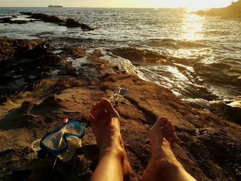 Low section of person relaxing on rock by sea during sunset