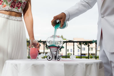 Midsection of bride and groom pouring mineral in glass container on table during wedding ceremony