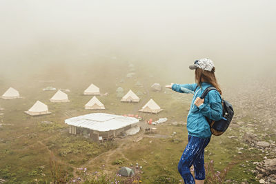 Young woman in cap with backpack hiker standing in mountain valley pointing hand at tent camping