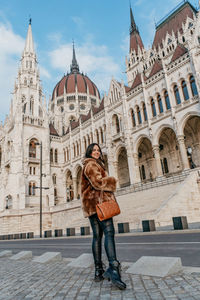 Young woman wearing stylish warm coat, standing in front of hungarian parliament in budapest hungary