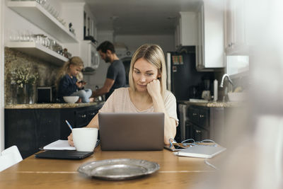 Mid adult woman using laptop at table with family in kitchen at home