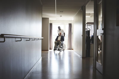 Full length of male nurse pushing senior man on wheelchair at hospital corridor